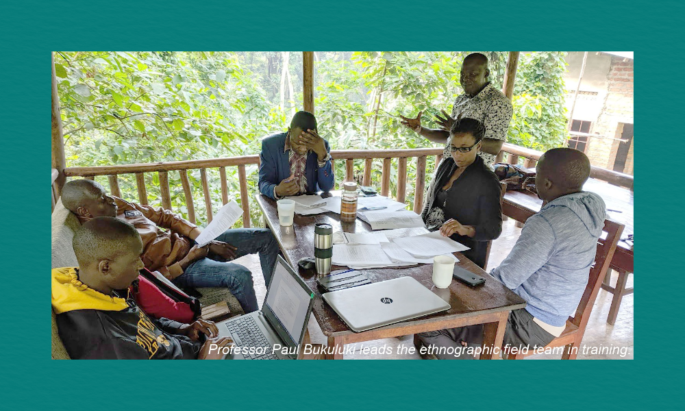 A small ethnographic field team sits around a wooden table on a covered porch surrounded by dense greenery. Papers, notebooks, coffee cups, and a laptop are spread across the table. One person stands and speaks while the others sit listening and reviewing documents. The scene conveys an informal outdoor training session led by Professor Paul Bukuluki.