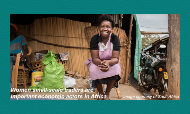 A woman sits smiling in front of a small wooden market stall surrounded by bags, baskets, and a nearby motorcycle, set within a local outdoor trading area.