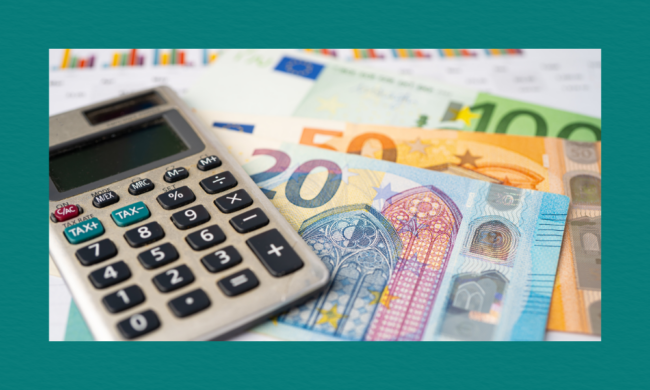 A close-up of a calculator resting on euro banknotes, with blurred colourful financial charts in the background.