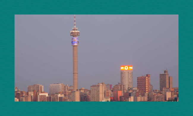 A city skyline at dusk with a tall telecommunications tower glowing blue near the top, surrounded by high‑rise buildings including one with a bright orange‑and‑white Vodacom sign, all under a clear, softly lit sky.