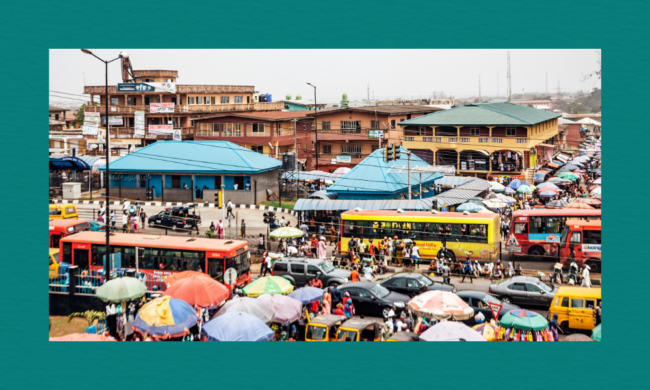 A crowded urban market with colourful umbrellas covering vendor stalls, tightly packed vehicles including buses, cars and motorcycles moving through heavy traffic, and people walking among multi‑storey buildings in the background.