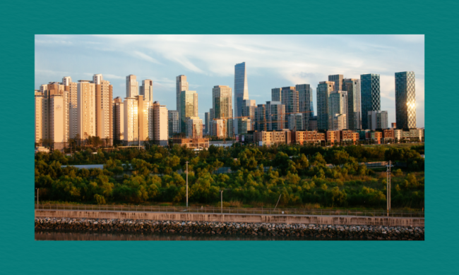 City skyline with modern high‑rise buildings and glass‑fronted towers behind a waterfront and a green area with trees, shown under a partly cloudy sky.