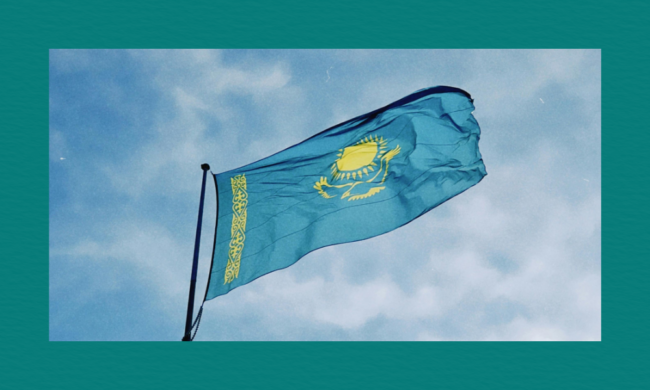 The Kazakhstan flag, light blue with a golden sun, eagle and vertical ornamental band, waves against a partly cloudy sky.