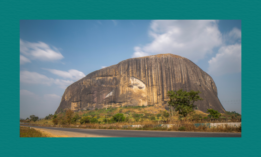Zuma Rock, a massive granite monolith with dark vertical streaks and a pale face‑like marking, rises beside a paved road in dry grassland under a partly cloudy sky.