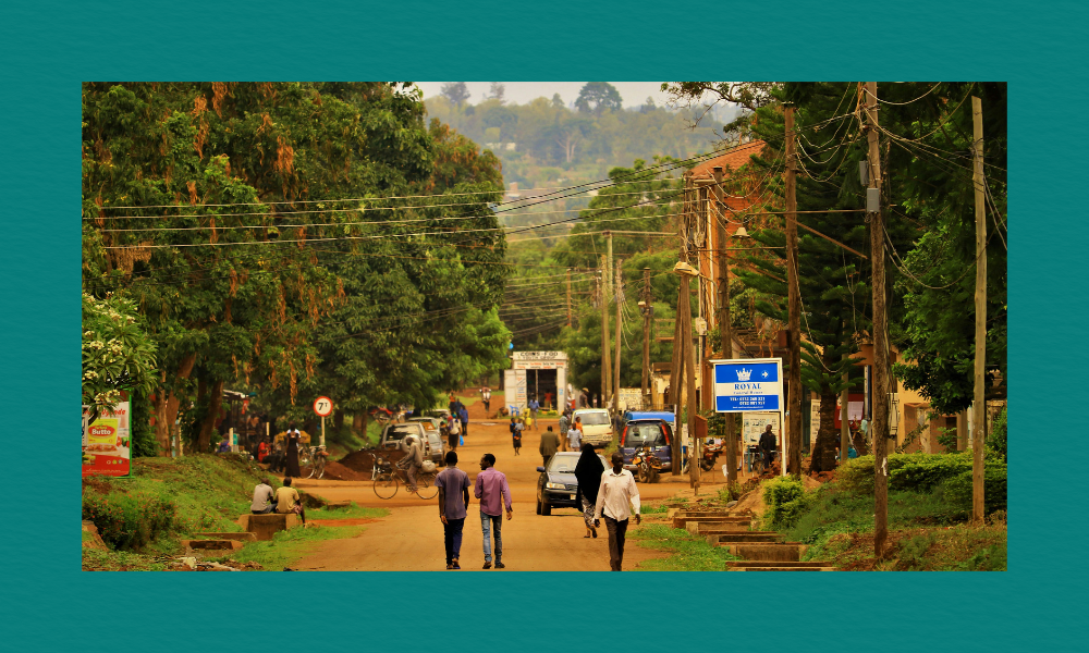 Unpaved street in a small town with people walking, cars and motorcycles along the road, lush greenery, and a visible sign for a hotel.