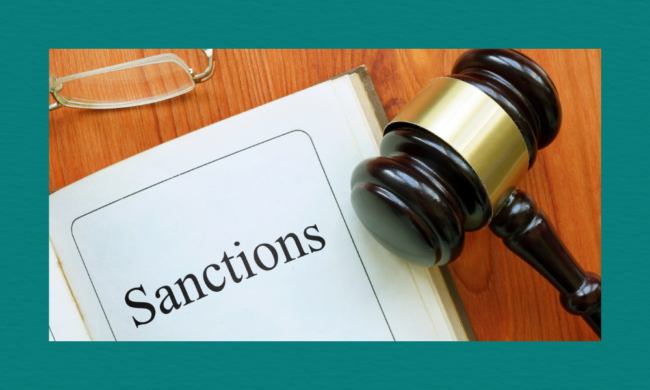 A wooden judge’s gavel rests on a polished wooden surface beside an open folder labeled “Sanctions” in bold black text, with a pair of eyeglasses placed near the top corner of the document.