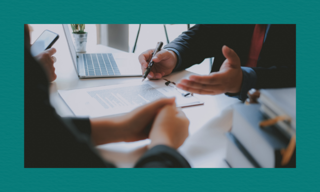Two people in business attire sit at a desk, reviewing a document on a clipboard beside an open laptop. One person holds a pen poised over the paper while the other gestures with a hand, suggesting discussion or decision‑making in a bright office setting.
