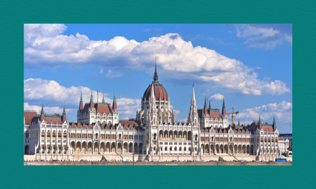 A wide daytime view of the Hungarian Parliament Building in Budapest, showing its ornate Neo‑Gothic façade, central dome, and spires along the Danube River under a bright blue sky with scattered clouds.