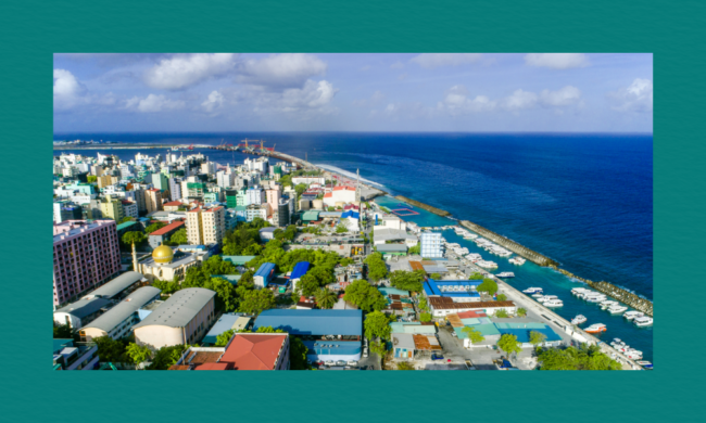 A high‑angle view of a densely built coastal city with colourful low‑rise buildings, a mosque with a golden dome, and a busy harbour lined with boats, all bordered by a long seawall and deep blue ocean under a partly cloudy sky.