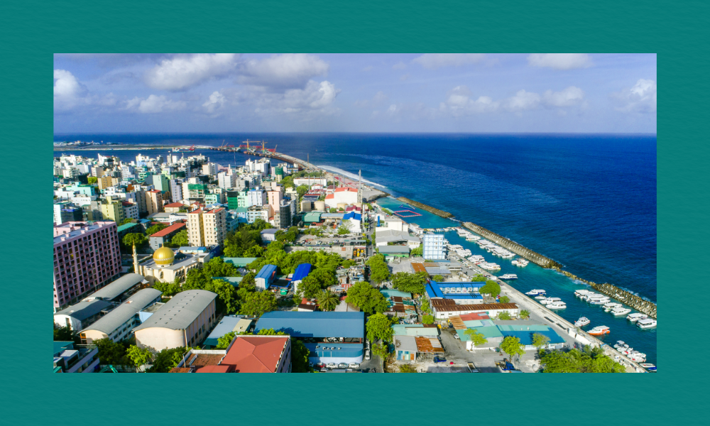 A high‑angle view of a densely built coastal city with colourful low‑rise buildings, a mosque with a golden dome, and a busy harbour lined with boats, all bordered by a long seawall and deep blue ocean under a partly cloudy sky.