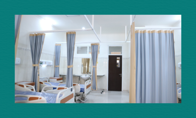 A clean, well‑lit hospital ward with several neatly arranged beds, each separated by light blue privacy curtains. The beds have blue mattresses and wooden accents, with small bedside tables beside them. Wall‑mounted medical equipment lines the tiled wall, and a dark wooden door and curtained window sit at the back of the room.