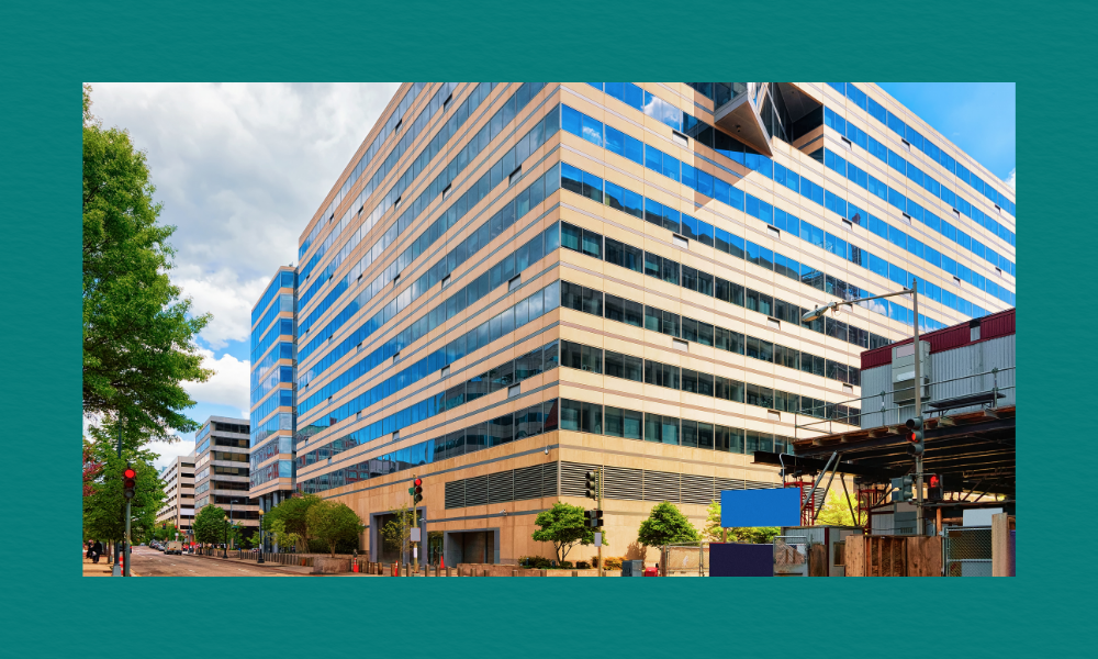 A modern multi‑storey glass and beige office building at a Washington, DC intersection, identified as the IMF headquarters. Reflective windows show the sky, while scaffolding and construction structures line the right side. Traffic lights, trees, and sidewalks frame the busy urban scene.