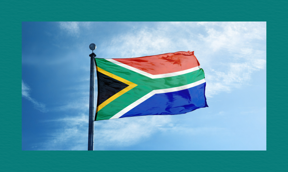 South Africa’s national flag flying on a tall flagpole against a bright blue sky with scattered white clouds. The multicoloured Y‑shaped design is clearly visible, with red, blue, green, black, yellow, and white sections.
