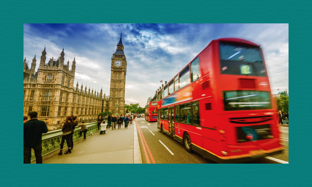 A busy London street scene featuring Big Ben and the Houses of Parliament on the left, with a red double‑decker bus driving across the bridge on the right. People walk along the pavement, some stopping to look at the view, under a partly cloudy blue sky.