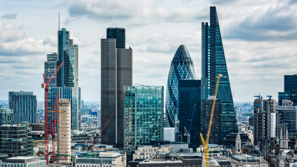 A view of London’s skyline featuring the Gherkin, the Cheesegrater, and other modern skyscrapers, with several construction cranes visible among the buildings. Older stone architecture sits in the foreground beneath a partly cloudy sky.