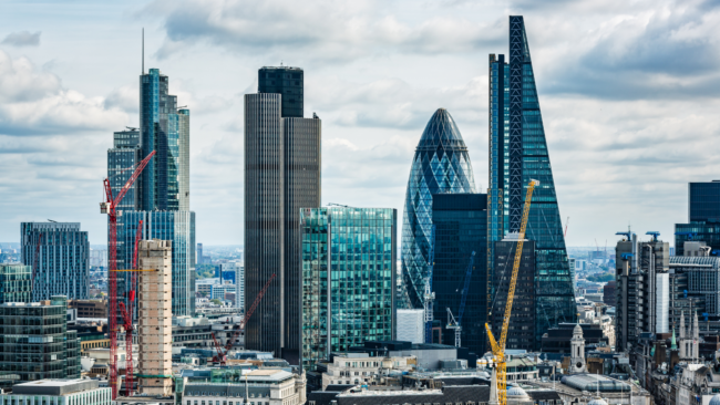 A view of London’s skyline featuring the Gherkin, the Cheesegrater, and other modern skyscrapers, with several construction cranes visible among the buildings. Older stone architecture sits in the foreground beneath a partly cloudy sky.