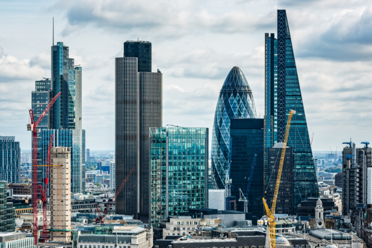 A view of London’s skyline featuring the Gherkin, the Cheesegrater, and other modern skyscrapers, with several construction cranes visible among the buildings. Older stone architecture sits in the foreground beneath a partly cloudy sky.