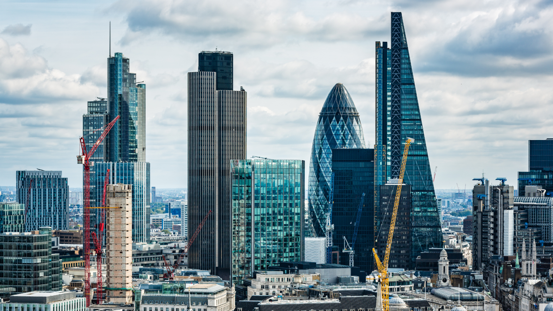 A view of London’s skyline featuring the Gherkin, the Cheesegrater, and other modern skyscrapers, with several construction cranes visible among the buildings. Older stone architecture sits in the foreground beneath a partly cloudy sky.