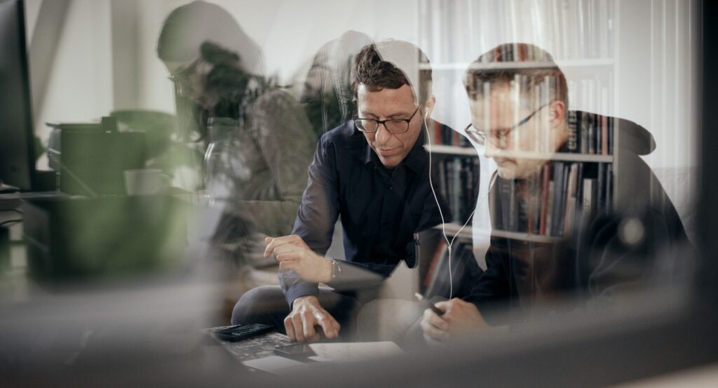 Two people sit at a desk in an office, viewed through reflective glass that overlays faint images of bookshelves and objects behind them. Both wear glasses and focus on documents or a device; one points at the desk while the other listens through earphones. The layered reflections create a complex, multi‑depth visual effect.