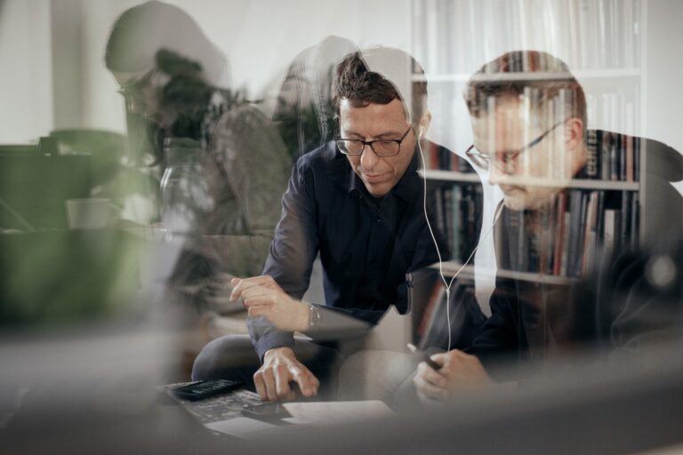 Two people sit at a desk in an office, viewed through reflective glass that overlays faint images of bookshelves and objects behind them. Both wear glasses and focus on documents or a device; one points at the desk while the other listens through earphones. The layered reflections create a complex, multi‑depth visual effect.