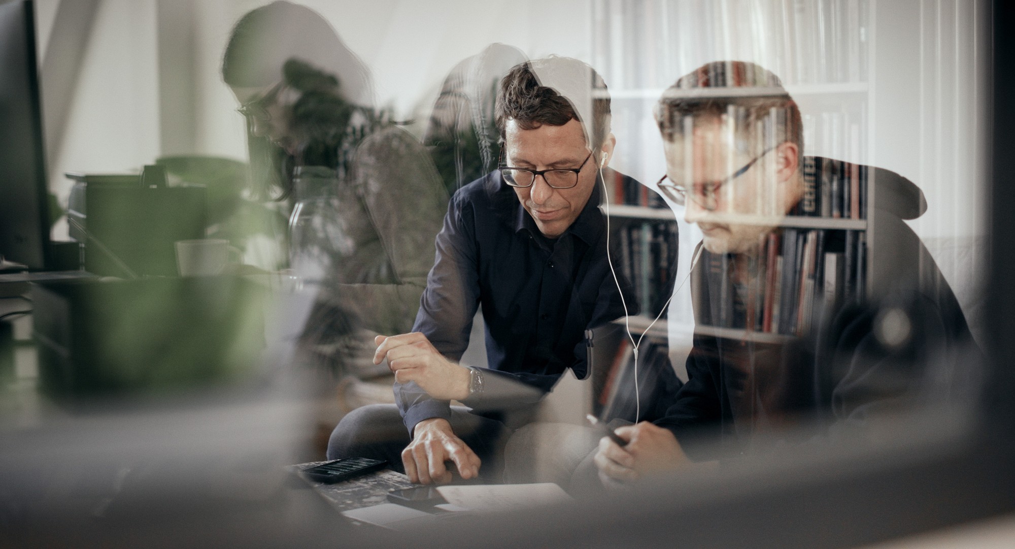 Two people sit at a desk in an office, viewed through reflective glass that overlays faint images of bookshelves and objects behind them. Both wear glasses and focus on documents or a device; one points at the desk while the other listens through earphones. The layered reflections create a complex, multi‑depth visual effect.