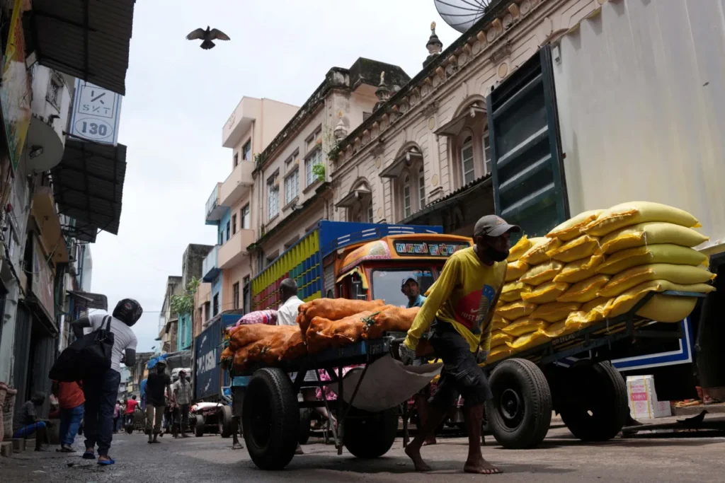 2025-03-18T120823Z_377656593_RC2UW9AQ7JT0_RTRMADP_3_SRI-LANKA-ECONOMY-GDP.jpg A narrow, bustling market street where workers pull heavy carts stacked with large orange and yellow sacks. A brightly painted truck is parked among weathered buildings with balconies, and a bird flies overhead. The scene conveys movement, labour, and the density of a busy commercial district.