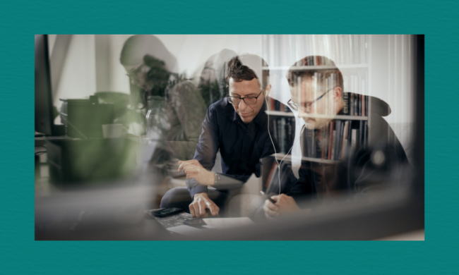 A new documentary shows the central role of whistleblowers in providing anti-corruption evidence A layered photographic composition showing two men with glasses working together at a desk, viewed through a reflective glass window. The reflection overlays multiple faint, translucent figures of other people working in an office environment, creating a double-exposure effect. In the foreground, one man points down at a document while the other holds a pen, both looking down intently under muted, dramatic lighting.