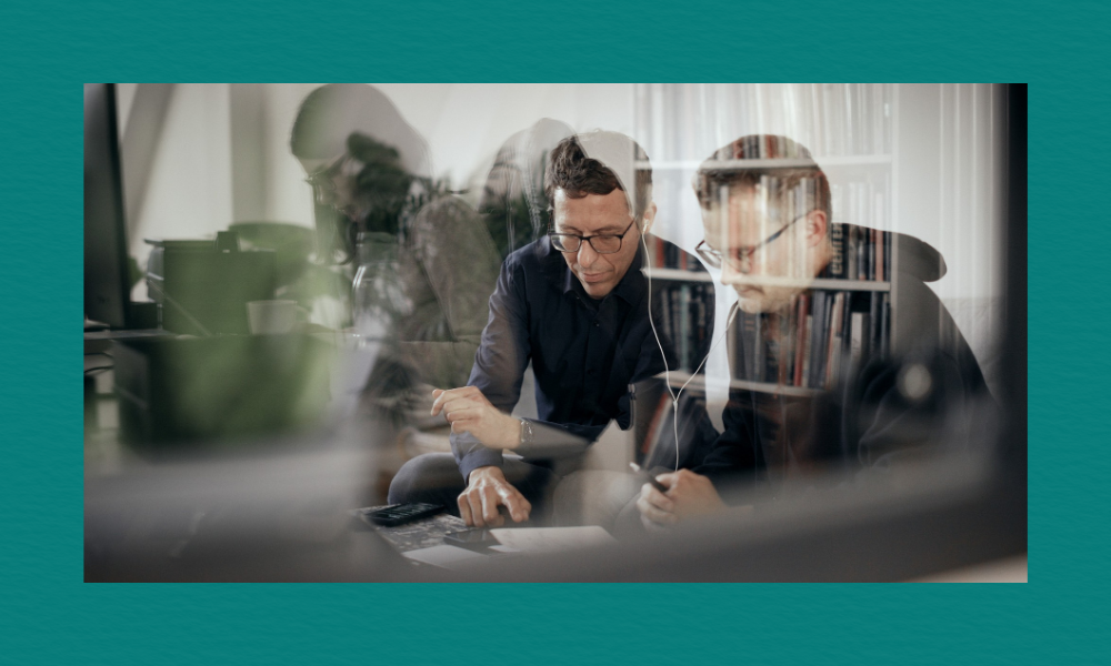 A layered photographic composition showing two men with glasses working together at a desk, viewed through a reflective glass window. The reflection overlays multiple faint, translucent figures of other people working in an office environment, creating a double-exposure effect. In the foreground, one man points down at a document while the other holds a pen, both looking down intently under muted, dramatic lighting.