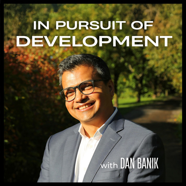 ab6765630000ba8ab1cf7773b1cf4aae9659f8d6 Portrait-style promotional image showing a man in glasses, a light blue shirt, and a blue blazer standing outdoors in bright sunlight, with trees and greenery behind him. Text at the top reads “In Pursuit of Development,” and at the bottom “with Dan Banik.”