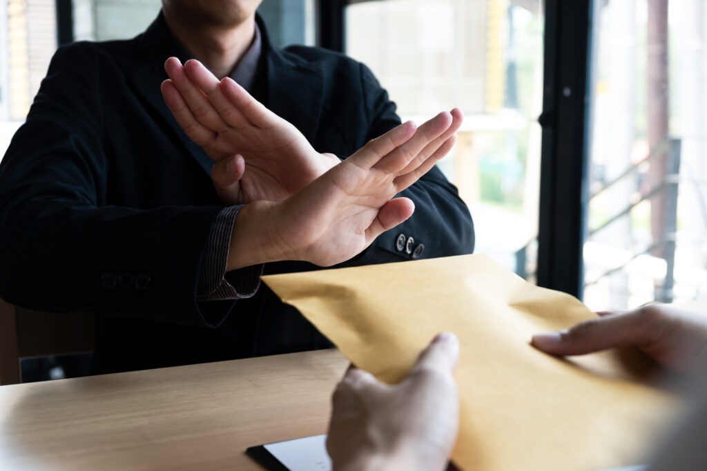 The businessmen refusing accept bribes in a brown envelope at the office. A person in a dark suit sits at a table with their arms crossed in an X‑shape across their chest, signalling refusal. Another person’s hands extend a large brown envelope toward them. The setting appears to be an office with windows in the background.
