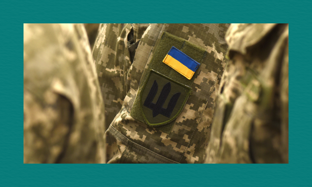 A close-up of a Ukrainian military uniform sleeve featuring two patches — a blue‑and‑yellow Ukrainian flag above a dark trident emblem, both sewn onto a camouflage fabric.