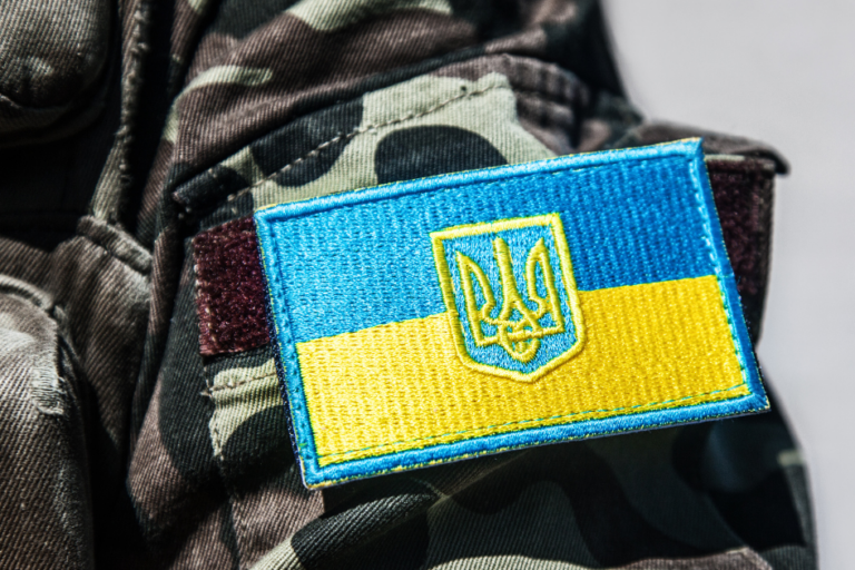 A close-up of a military uniform in camouflage fabric featuring a sewn patch of the Ukrainian flag with a blue-and-yellow design and a gold trident emblem in the centre.