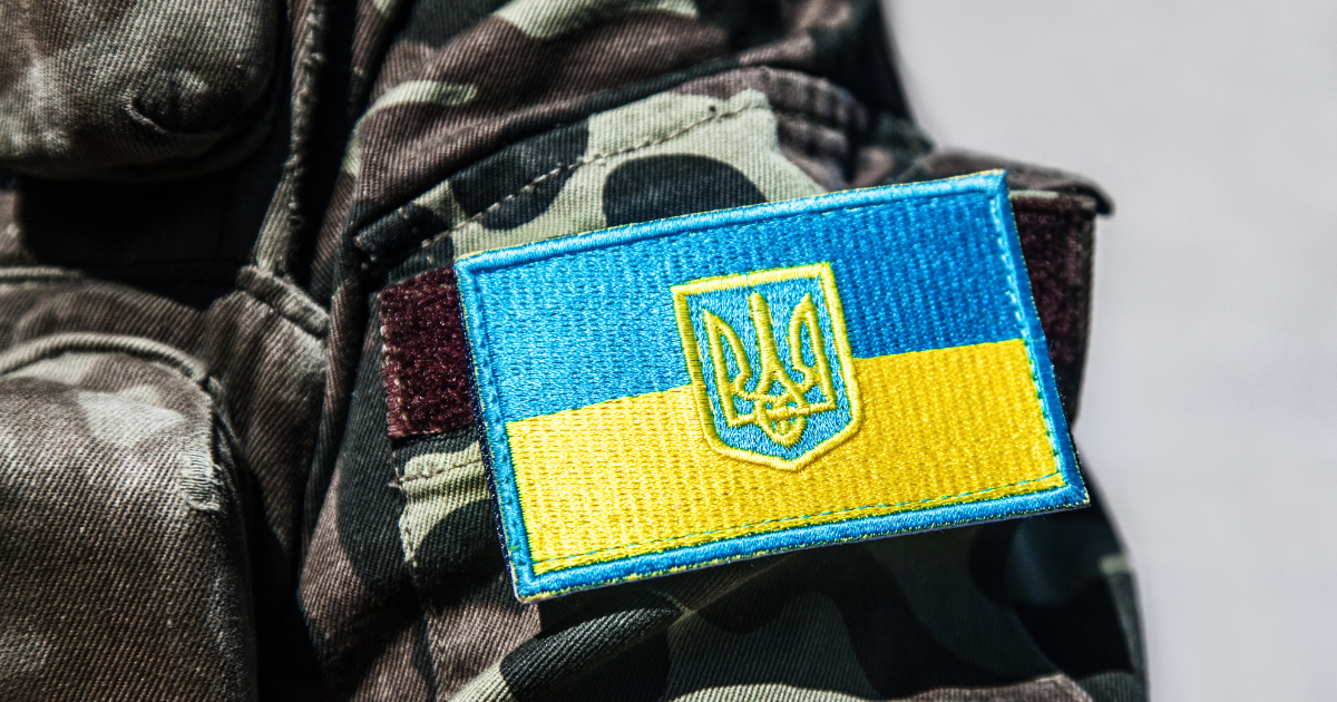 A close-up of a military uniform in camouflage fabric featuring a sewn patch of the Ukrainian flag with a blue-and-yellow design and a gold trident emblem in the centre.