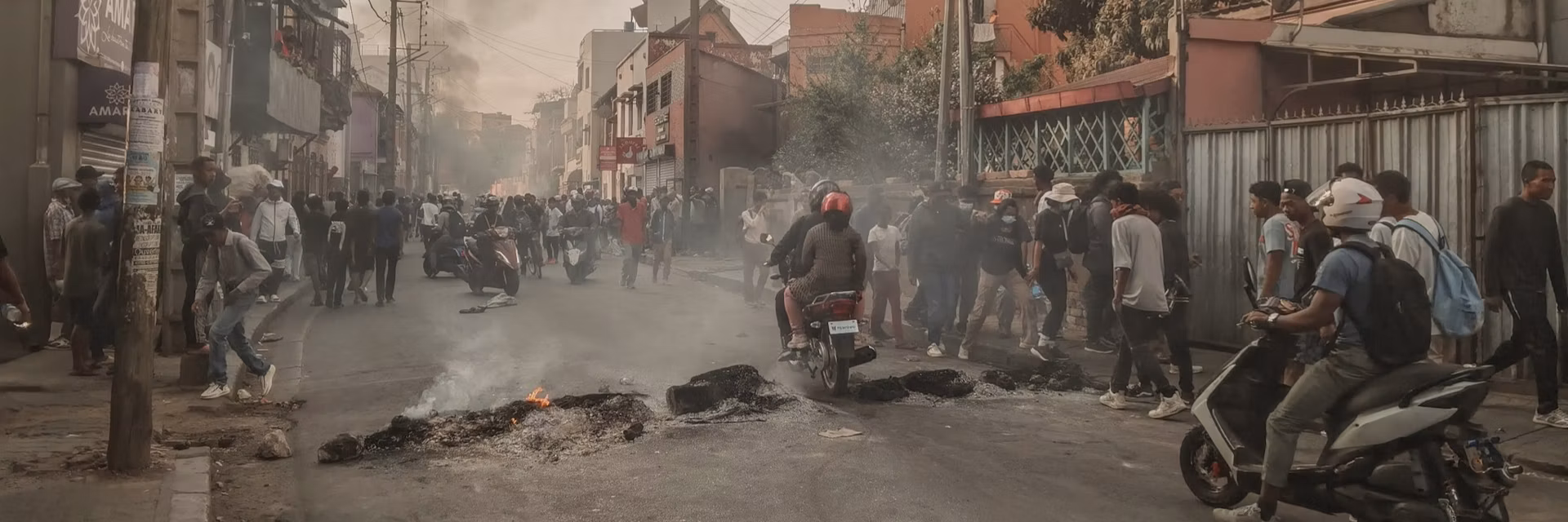 Crowded street in Madagascar during a protest, with people gathered around burned debris and smoke rising from the road. Two motorcyclists wearing helmets ride through the scene while groups of onlookers stand near buildings lining the street.