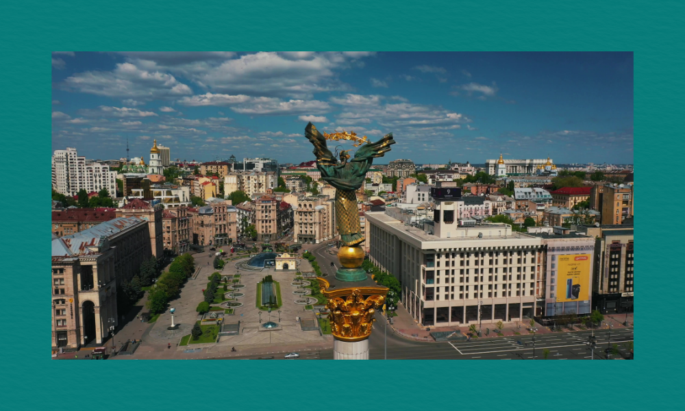 View of Freedom Square in Kyiv, Ukraine, featuring the tall Independence Monument topped with a bronze figure holding a branch. Surrounding the square are historic and modern buildings, with golden domes visible in the background. Fountains, landscaped areas, and traffic line the streets under a bright, partly cloudy sky.