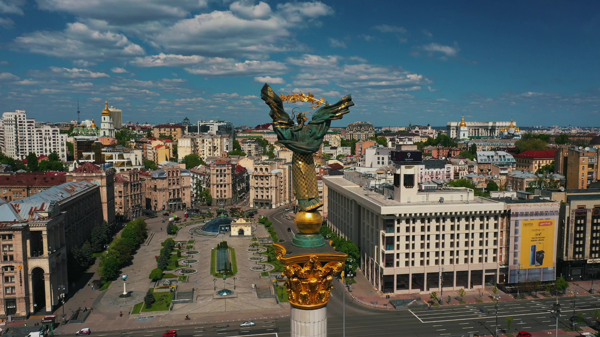 View of Freedom Square in Kyiv, Ukraine, featuring the tall Independence Monument topped with a bronze figure holding a branch. Surrounding the square are historic and modern buildings, with golden domes visible in the background. Fountains, landscaped areas, and traffic line the streets under a bright, partly cloudy sky.